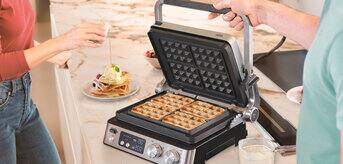 Person making waffles on a kitchen counter while another person adds syrup to plated waffles.
