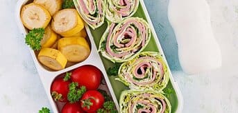 Lunchbox z rolkami szynkowymi, bananami, pomidorami i napojem w butelce obok. - zdjęcie