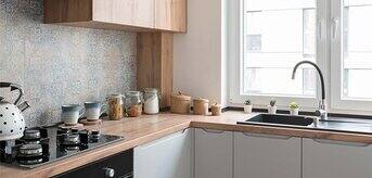 Nowoczesna kuchnia z drewnianymi akcentami, jasnymi frontami szafek i dużym oknem. Widać strefę gotowania z płytą grzewczą i piekarnikiem oraz zlewozmywak z roślinami na parapecie. - zdjęcie