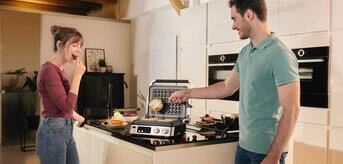 A couple standing in their kitchen preparing waffles in a Braun multifunctional contact grill.