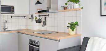 Nowoczesna, mała kuchnia z szarymi meblami, drewnianym blatem, białymi kafelkami i roślinami w doniczkach. W tle widoczna jest część przestronnego salonu z szarą kanapą. - zdjęcie