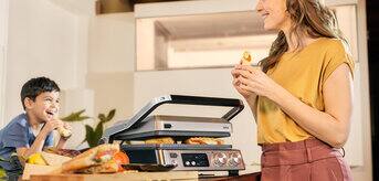 A woman and a boy enjoy slices of pizza together while seated in front of Braun's MultiGrill 7.