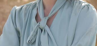 Kobieta w jasnozielonej bluzce z jedwabiu, z wiązaniem na kokardę przy szyi, sfotografowana na zewnątrz. - tytułowa