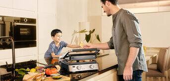 A man and a child cook food in a kitchen using Braun MultiGrill 7. The child hands something to the man.