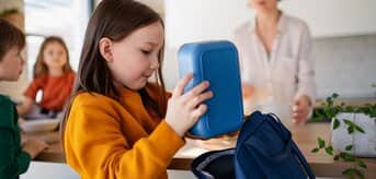 Dziecko pakujące lunch do plecaka, podczas gdy dorosły stoi w tle. - tytułowa