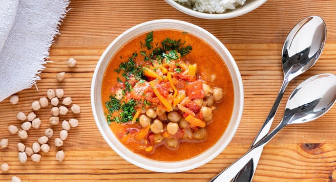 Top view of a wooden table with a soup plate with chickpea coconut curry and a bowl of rice.