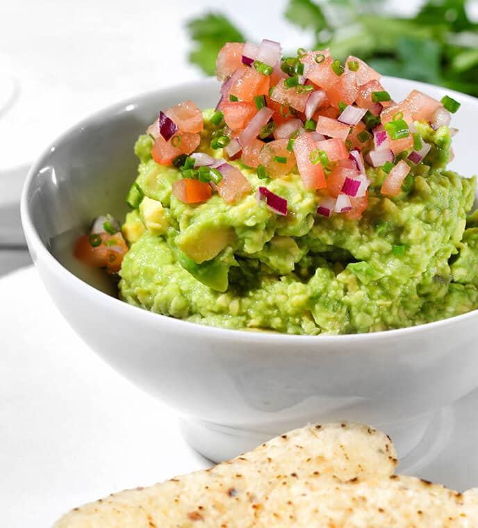 A white bowl of guacamole with a topping of diced tomatoes and onions